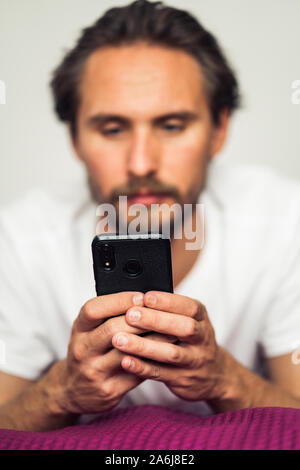 Foto di un piuttosto grave giovane uomo bello interagire con il suo dispositivo mobile mentre giaceva a letto Foto Stock