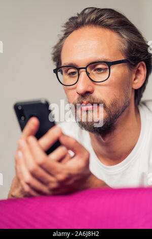 Foto di un piuttosto grave giovane uomo bello interagire con il suo dispositivo mobile mentre giaceva a letto Foto Stock