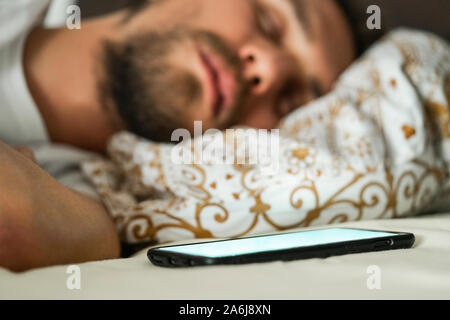 Close-up foto di un giovane uomo dorme nel letto. Telefono cellulare con un bianco schermo lampeggiante è visibile in primo piano. Foto Stock