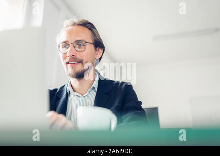 Foto di un bello concentrato e sorridente giovane uomo seduto con la scrivania a lavorare al computer. Tazza di caffè visibile in primo piano. Foto Stock