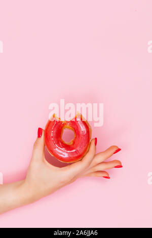 La mano di una bella donna detiene un rosso dolce morso ciambella con bagliori. Primo piano delle mani di una giovane donna con manicure sulle unghie. Foto Stock