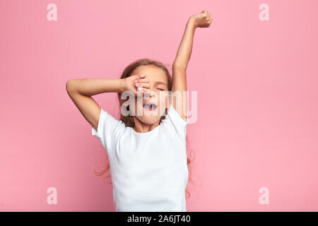 Ragazza carina bambino con lunghi capelli biondi sbadigli dolcemente, allungando le braccia, kid vuole dormire, isolato backround rosa, stanchezza. close up verticale Foto Stock