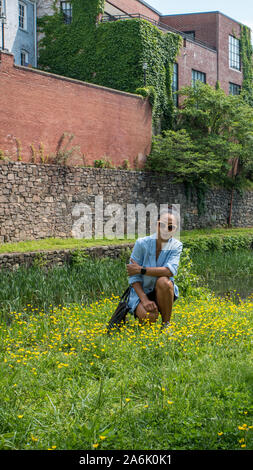 Donna con occhiali da sole in ginocchio in erba accanto al canal in montagna con edifici in mattoni in background Foto Stock