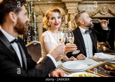 Elegantemente vestito gruppo di persone aventi una cena di gala in una tavola ben servita vicino all'albero di natale, per celebrare il nuovo anno di vacanza presso il ristorante di lusso Foto Stock