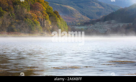 Nebbia rotolamento sulla superficie del lago sulla soleggiata mattina autunnale nelle highlands scozzesi.bella scena di paesaggio con umore atmosferica.Loch e collina con alberi. Foto Stock