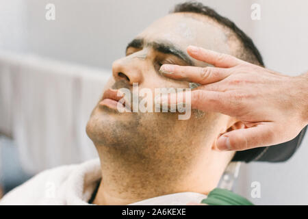 La pelle maschile cura nel salone di bellezza. Applicazione di argilla Maschera di pulizia su una il volto dell'uomo Foto Stock