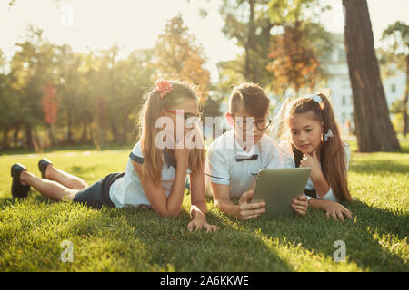 Tre sorridente in età scolastica sono amici sdraiati sull'erba e guardando il tablet Foto Stock