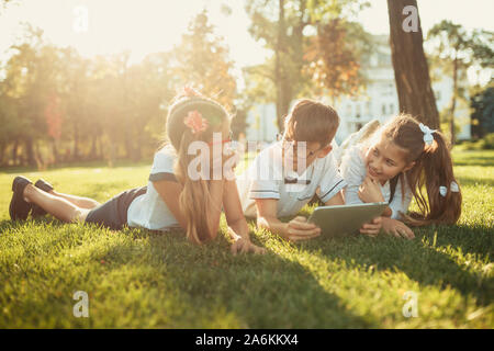 Tre sorridente in età scolastica sono amici sdraiati sull'erba e guardando il tablet Foto Stock
