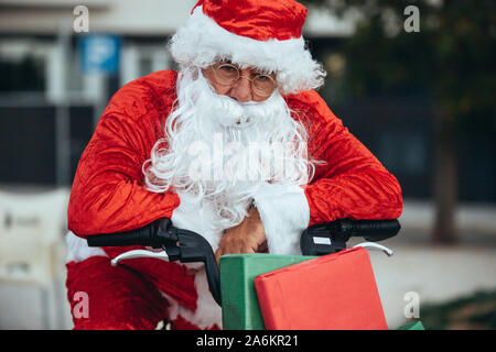 Magazzino verticale foto di santa claus supportato in una bicicletta piena di regali con espressione esausta. Il tempo di Natale Foto Stock