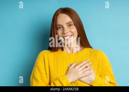 Bella grati amichevole-cercando redhead caucasian donna con sorriso affascinante in maglione giallo mantenendo entrambe le mani sul petto, esprimendo gratitudine,sh Foto Stock