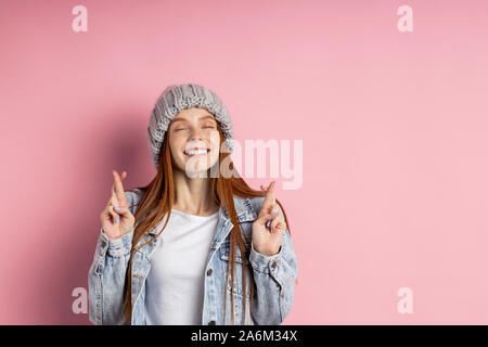 Felice lieto redhead donna in camicia di denim in posa con le dita incrociate e gli occhi chiusi, pregando per la buona fortuna sorride isolate su sfondo blu. Foto Stock