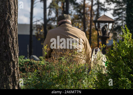 Un vecchio uomo grigio in uno strato leggero e hat piange presso la sua tomba passante in un cimitero in autunno in una giornata di sole. Un cestello posto a fianco di un uomo. Foto Stock