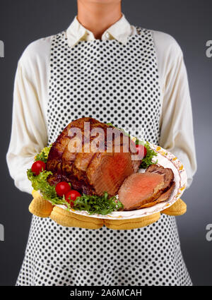 Primo piano di una massaia in un grembiule e guanti da forno tenendo un piatto di arrosto di manzo, tagliato a fette con guarnire. Foto Stock