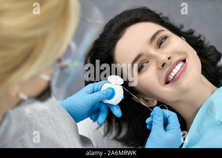 Felice ragazza in un paziente in bib una clinica dentale. Dentista di guanti in lattice blu si prepara a filo interdentale i suoi denti con un aiuto di un filo interdentale. Primo piano. H Foto Stock