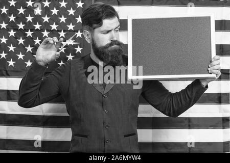 Educazione patriottica. Uomo Barbuto holding forbici e Blackboard nella scuola di educazione. Barbiere corsi di istruzione. Hipster dando lezione sulla bandiera americana sfondo. Istruzione e carriera, copia dello spazio. Foto Stock