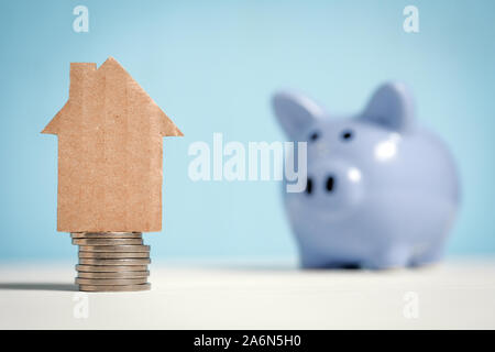 Abstract casa di cartone su una pila di monete accanto a un salvadanaio. Immobiliare di concetto di risparmio. Close up. Foto Stock