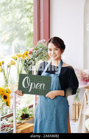 Bella sorridente fiorista vietnamita in grembiule blu tenendo aperte segno e guardando la fotocamera Foto Stock