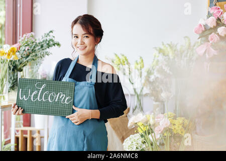 Piuttosto giovane vietnamita fioraio azienda cartello di benvenuto e accoglie i clienti per il suo negozio di fiori Foto Stock