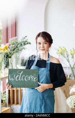 Ritratto di giovane donna asiatica in azienda i grembiuli di cartello di benvenuto e invitando il cliente per il suo negozio di fiori Foto Stock