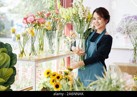 Sorridente giovane donna asiatica immissione vasi con vari splendidi fiori che sbocciano in fiore boutique Foto Stock