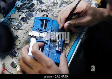 Repairman utilizzando le pinzette per applicare o rimuovere la pasta di saldatura sulla scheda logica dello smartphone Foto Stock