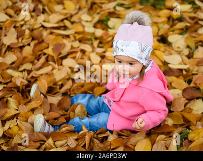 Baby girl in autunno park seduto in mucchio di foglie secche avendo divertimento. Tema di autunno Foto Stock