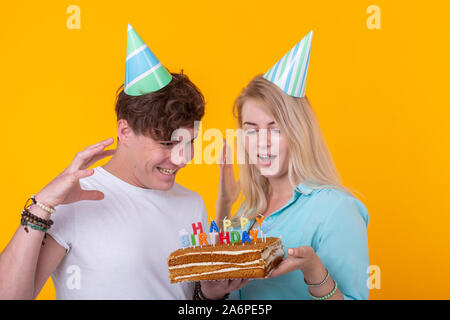 Divertente coppia giovane di tappi di carta e con una torta a fare una faccia stolto e auguro buon compleanno mentre in piedi contro uno sfondo giallo. Concetto di Foto Stock