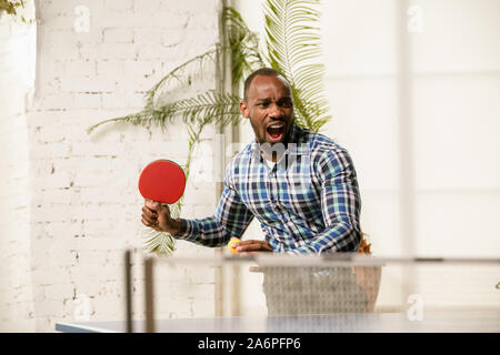 Giovane uomo giocando a ping-pong nel luogo di lavoro, avendo divertimento. Modello in abiti casual giochi ping pong eccitante al giorno di sole. Concetto di attività per il tempo libero, sport, amicizia, teambuilding, il lavoro di squadra, emozioni. Foto Stock