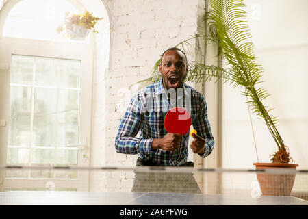 Giovane uomo giocando a ping-pong nel luogo di lavoro, avendo divertimento. Modello in abiti casual giochi ping pong eccitante al giorno di sole. Concetto di attività per il tempo libero, sport, amicizia, teambuilding, il lavoro di squadra, emozioni. Foto Stock