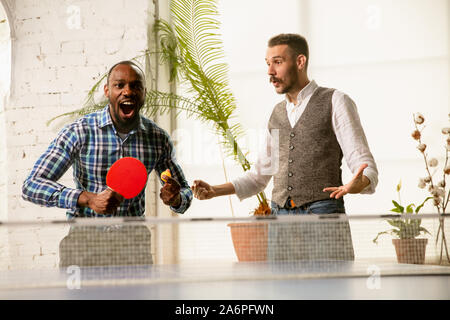 Giovani uomini giocando a ping-pong nel luogo di lavoro, avendo divertimento. Amici in abiti casual giocare a ping pong insieme alla giornata di sole. Concetto di attività per il tempo libero, sport, amicizia, teambuilding, il lavoro di squadra. Foto Stock