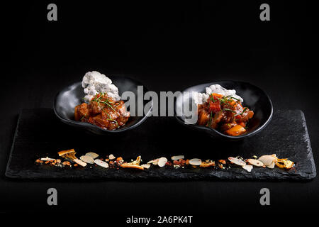 Primo piano sul gustoso stufato con ortaggi: patate, carote, pepe rosso, cosparsa di prezzemolo, servita in piastre nere con un cucchiaio al buio su un tavolo. Foto Stock