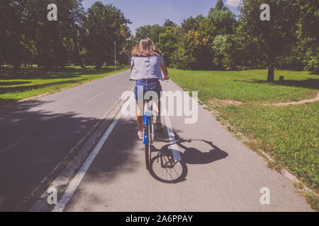 Donna graziosa bicicletta equitazione nel Parco al giorno di estate , i jeans corti , vista posteriore. Foto Stock