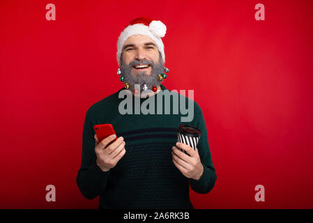 Felice l'uomo barbuto che indossa santa claus hat tenendo lo smartphone e tazza di caffè per andare Foto Stock