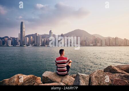 Giovane uomo seduto sulle pietre contro Hong Kong cityscape con grattacieli al tramonto colorato. Foto Stock
