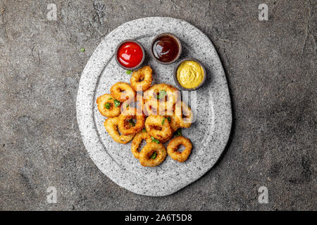 Calamari fritti anelli in pietra grigia piastra con salse. Grigio sfondo concreto Foto Stock