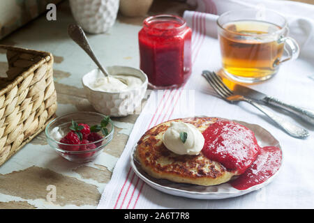 Frittelle al curdo con salsa di fragole, panna acida e lamponi e tè alle erbe su un tavolo leggero. Stile rustico. Foto Stock
