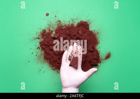 Pezzo di cioccolato tenuto in mano con guanto sulla pila di polvere di cacao, su sfondo verde. Contesto di pasticceria. La produzione del cioccolato. Ingrediente di dessert Foto Stock