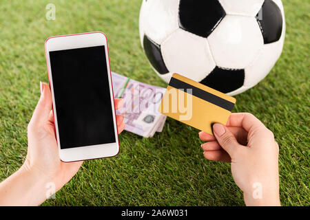 Vista ritagliata della mani femminili con lo smartphone e carta di credito vicino al pallone da calcio e le banconote in euro sulla verde erba, scommesse sportive concept Foto Stock