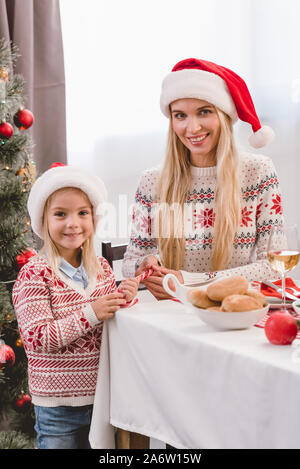 Madre e figlia in maglioni e cappelli di Babbo Natale sorridente e guardando la telecamera nel Natale Foto Stock