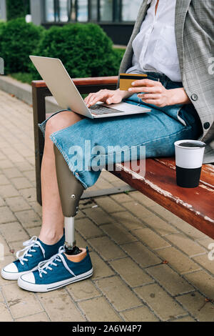 Vista parziale di donna disabile con laptop seduta sul banco di lavoro e tenendo carta di credito Foto Stock