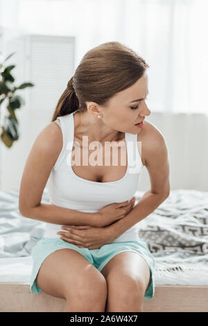 Sconvolto giovane donna seduta sul letto e toccando la pancia Foto Stock