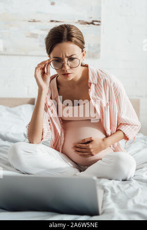 Preoccupato per la donna incinta in bicchieri seduta sul letto e utilizzando computer portatile Foto Stock