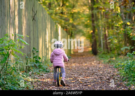Giovane ragazza in abiti caldi in equilibrio in bicicletta lungo un sentiero autunnale oltre ad una alta recinzione di legno nella foresta. Visto in ottobre in Germania, B Foto Stock