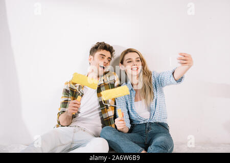 Allegro coppia giovane tenendo selfie mentre è seduto vicino a muro bianco e giallo di contenimento i rulli di verniciatura Foto Stock