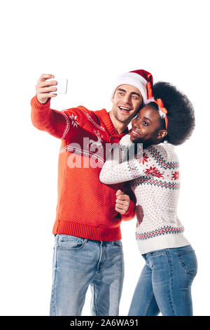 Coppia multiculturale in santa hat e corna di cervo tenendo selfie sullo smartphone, isolato su bianco Foto Stock