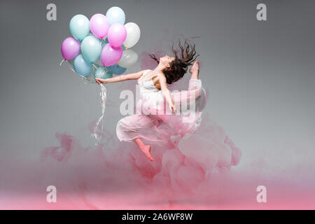 Bellissima ballerina ballando con palloncini di festa nei pressi di rosa gli spruzzi di fumo su sfondo grigio Foto Stock