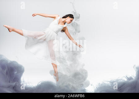 Giovane ballerina leggiadra in abito bianco dancing in fumo grigio su sfondo grigio Foto Stock