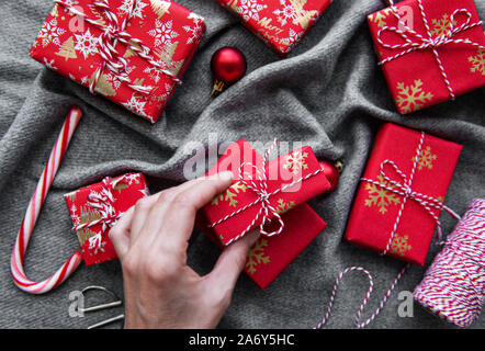 Sfondo di natale con confezioni regalo, bugne di corda e decorazioni su rosso. Preparazione per le vacanze. Vista dall'alto. Donna di mani confezione regalo. Foto Stock