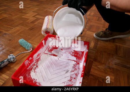 Lavoratore edile e tuttofare finitura facendo lavori di rinnovo nell'appartamento. Pittore professionista con guanti riversa il colore bianco dal cucchiaio nel recipiente rosso. Foto Stock