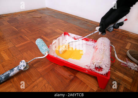 Lavoratore edile e pittore con guanti contiene spazzola a rullo la vernice in mano e mix di vernice gialla nel contenitore e rosso ciotola per miscelare i colori. Foto Stock